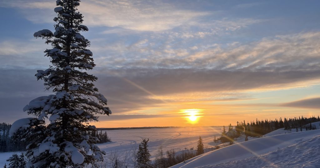 A sunset or sunrise scene over a snow covered landscape with a single fir tree in the background.